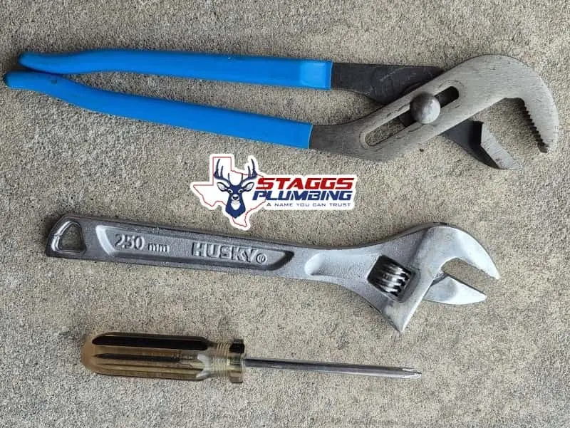 Plumbing tools laid out for an outdoor spigot repair, including an adjustable wrench, channel lock pliers, screwdriver, and water meter key. Staggs Plumbing shows the basic toolkit needed to fix a leaky hose bibb at home.